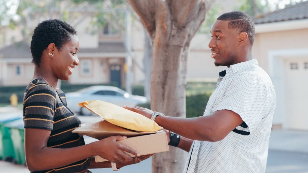 A courier delivers packages to a smiling woman at an outdoor location, representing online shopping and delivery services.
